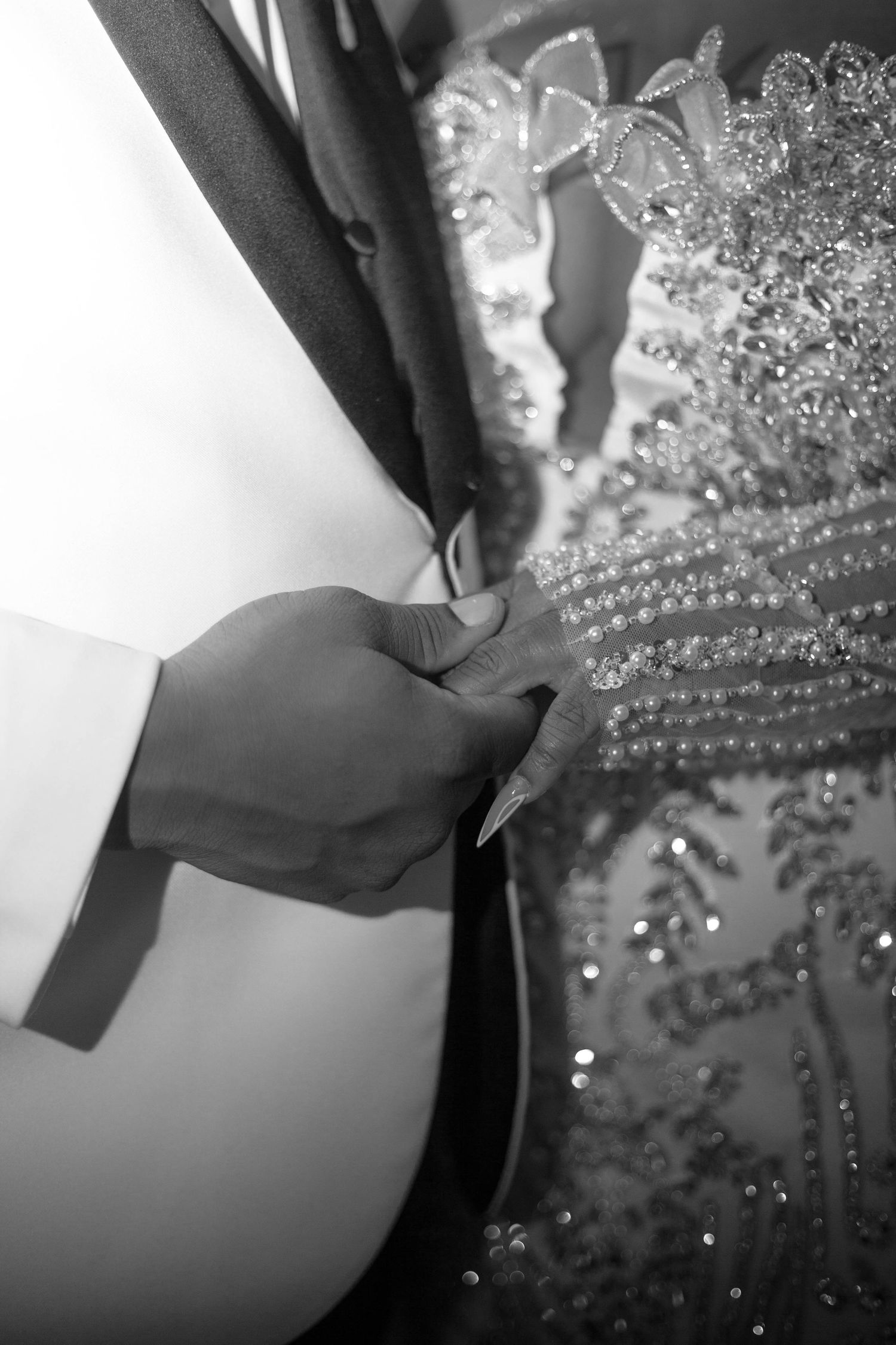 Black and white close-up of a groom in a white tuxedo jacket holding the hand of a bride in a beaded, sequined gown.