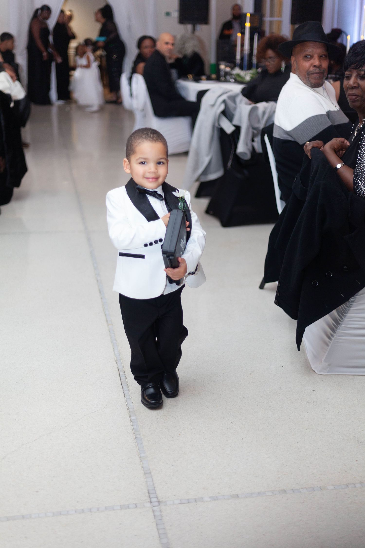 Young ring bearer in a white tuxedo jacket and black bow tie walks across a wedding reception floor while guests sit at tables behind him.