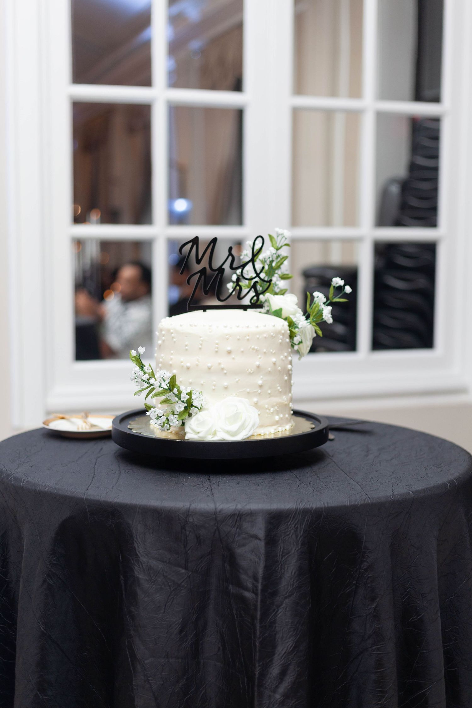 White pearl-beaded wedding cake with white roses, greenery and a black Mr & Mrs topper on a black draped table by a window.