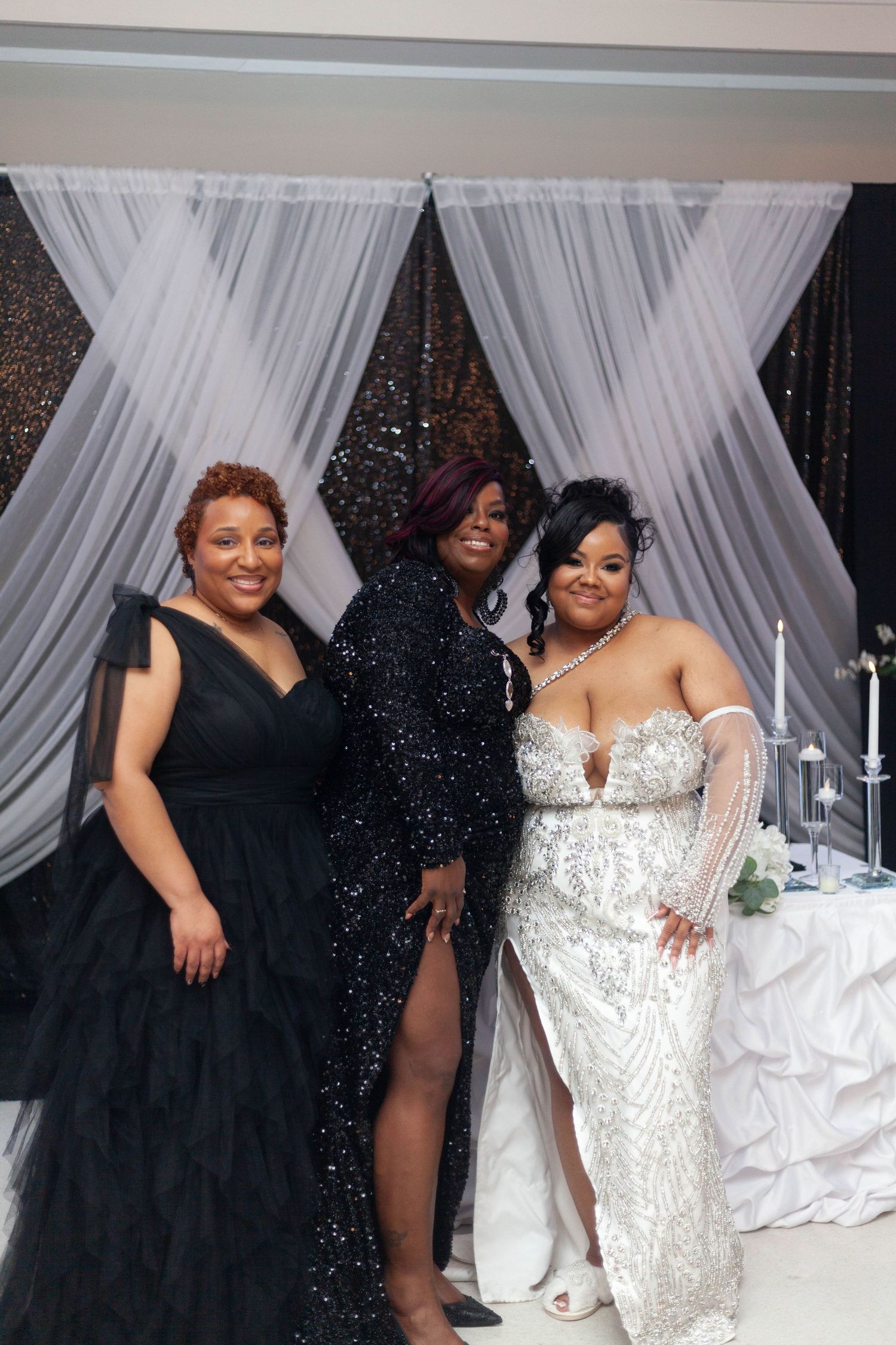 Three women pose together at a wedding reception, two in black gowns and one in a sparkling white gown, beside a table with candles.