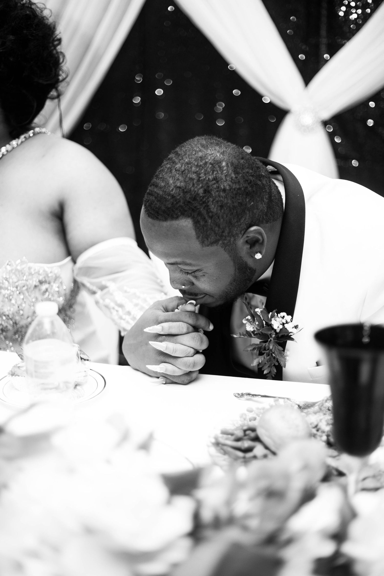 Groom in a white tuxedo jacket with a floral boutonniere bows his head in prayer at a wedding reception table beside the bride.
