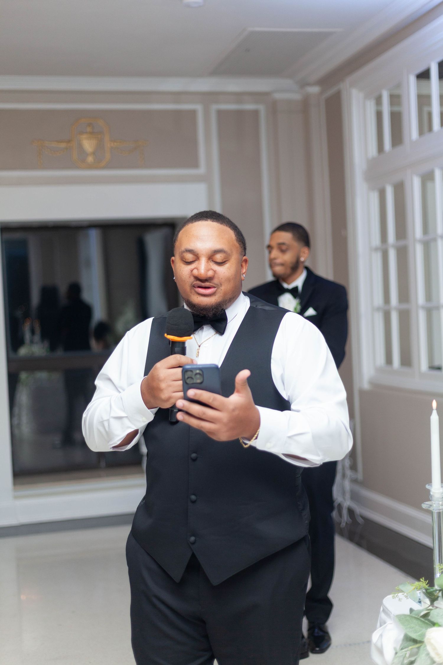 A man in a black vest and bow tie speaks into a microphone while holding a phone at an elegant formal event, with another man behind him.