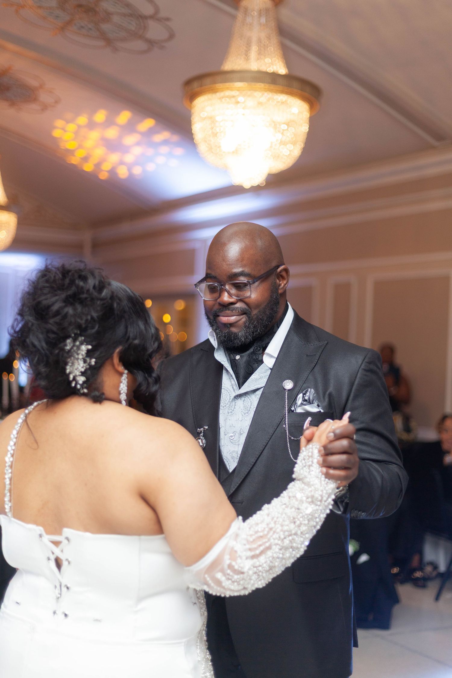 A bride in a white lace gown and a groom in a gray suit share a first dance under a crystal chandelier at an elegant wedding reception.