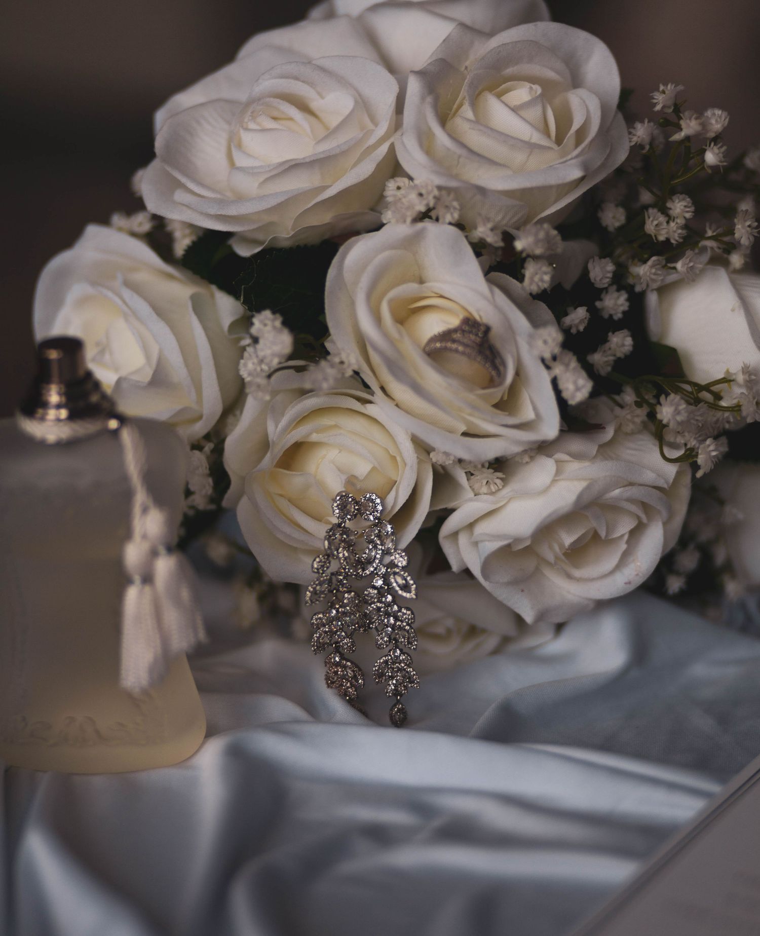 White rose bridal bouquet with baby's breath beside sparkling crystal drop earrings and a perfume bottle on soft satin fabric.