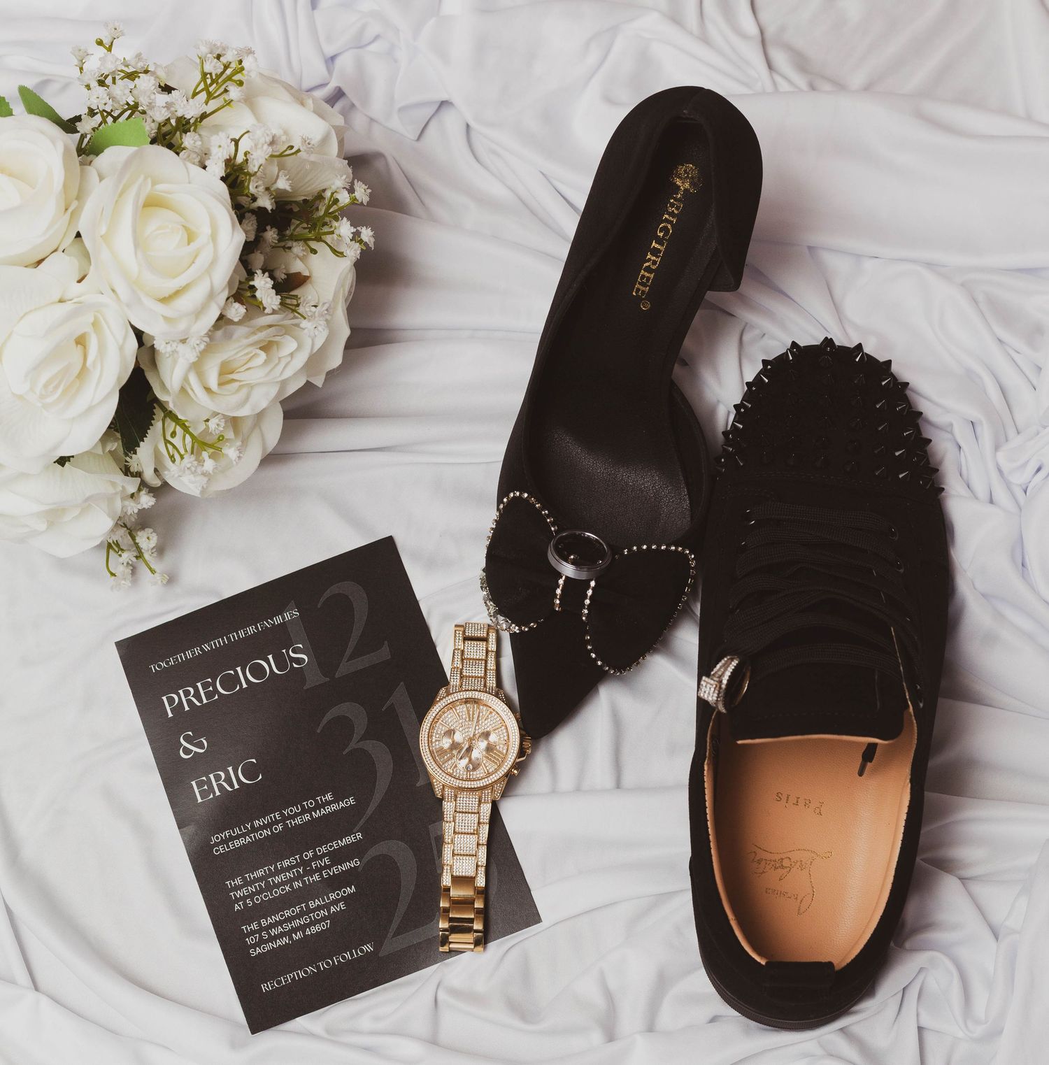 Black wedding invitation for Precious and Eric with a gold watch, black bow heel, spiked loafer and white rose bouquet on satin.
