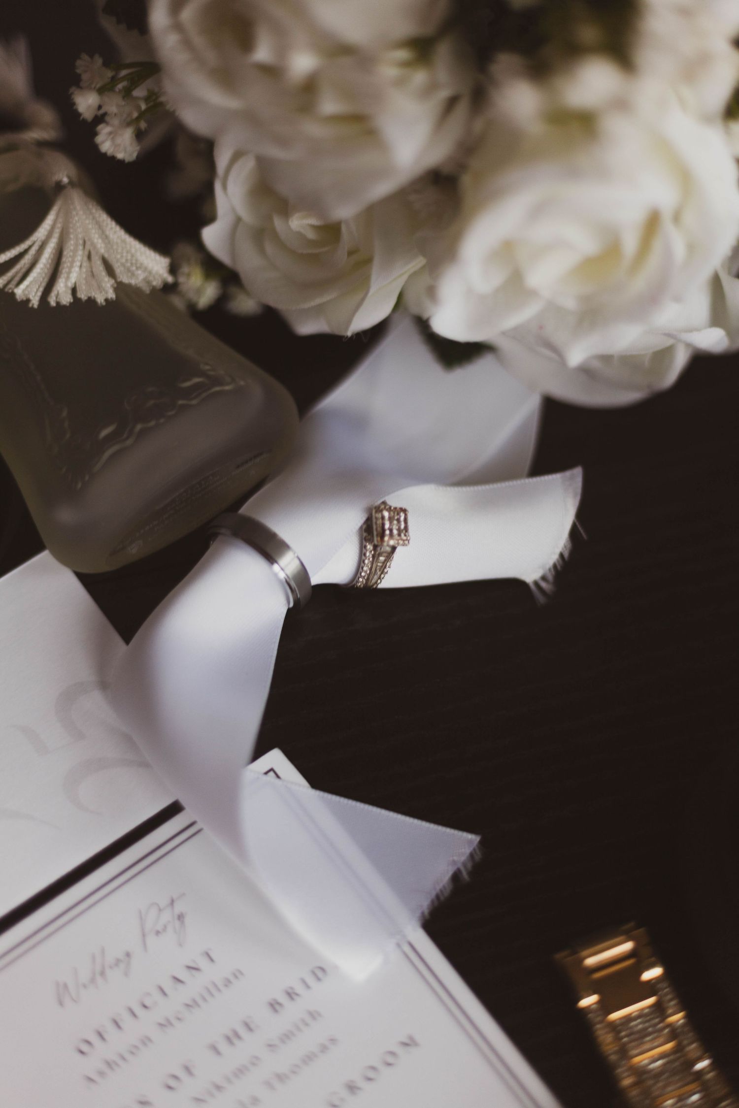Wedding rings and a gold watch rest on a white silk ribbon beside white roses and a printed wedding party program.