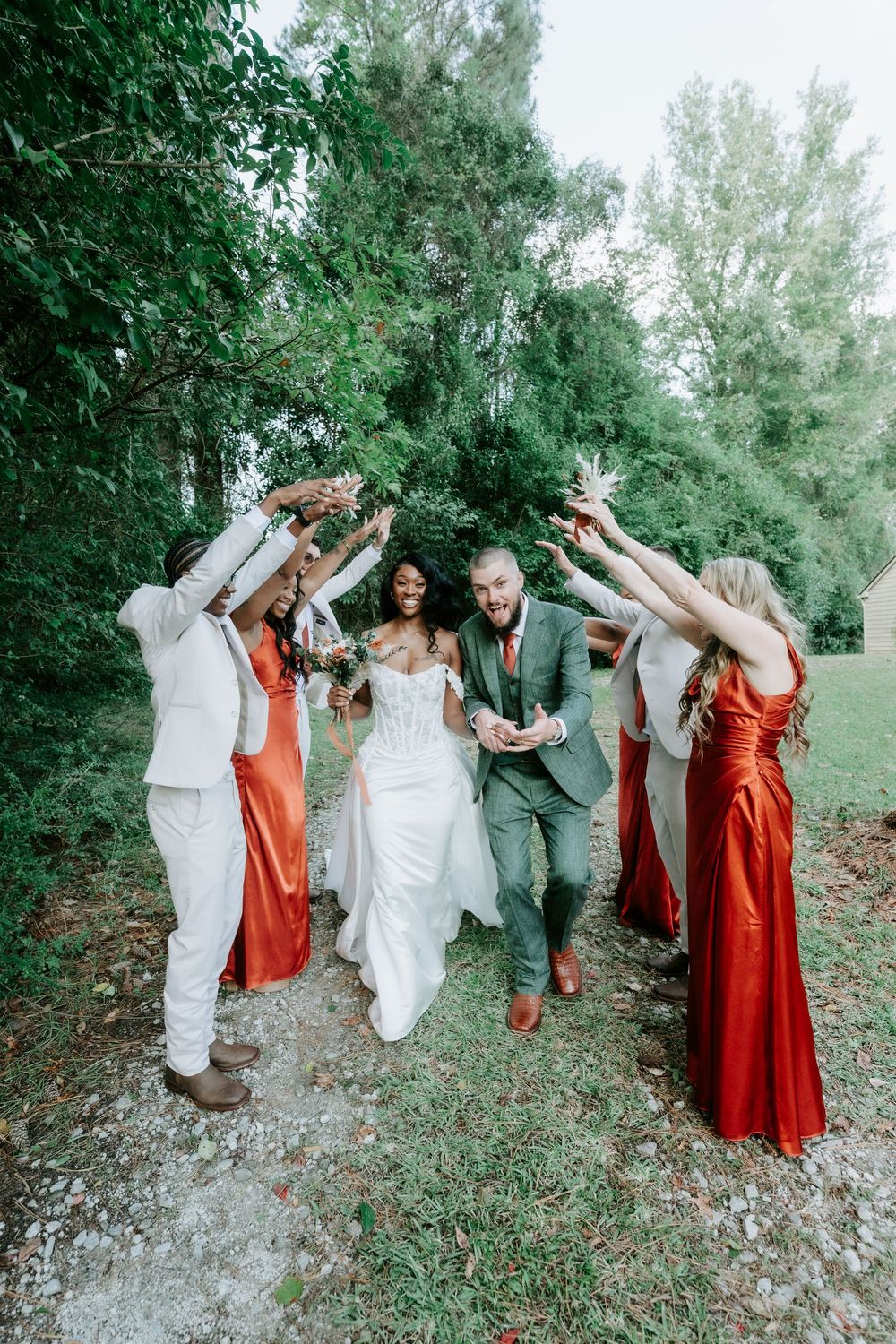 Bride and groom walk hand in hand through an arch of raised bouquets formed by bridesmaids in rust dresses and groomsmen in cream suits.