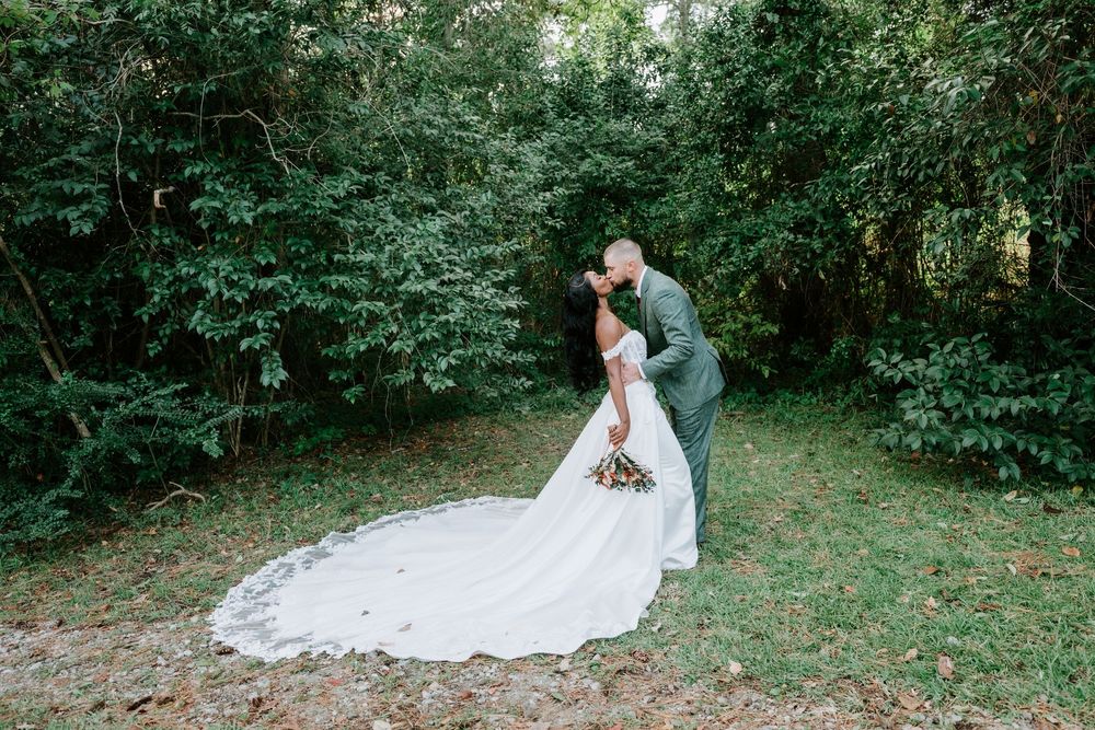 A bride in a long white lace gown and a groom in a green suit share a kiss on a grassy path surrounded by lush green trees.