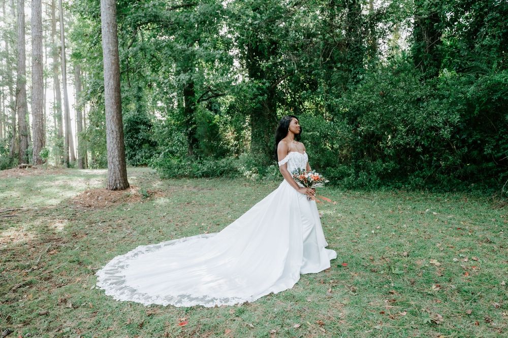 Bride in an off-the-shoulder white wedding gown with a long lace-trimmed train holds a bouquet on a grassy lawn surrounded by tall trees.