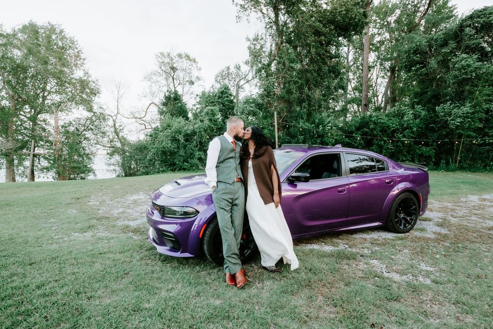 Couple embracing and kissing beside a purple Dodge Charger on a grassy lawn surrounded by tall green trees.