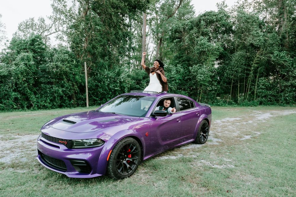 Two women pose with a purple widebody Dodge Charger on a grassy field, one leaning out the driver window and one standing on the door frame.