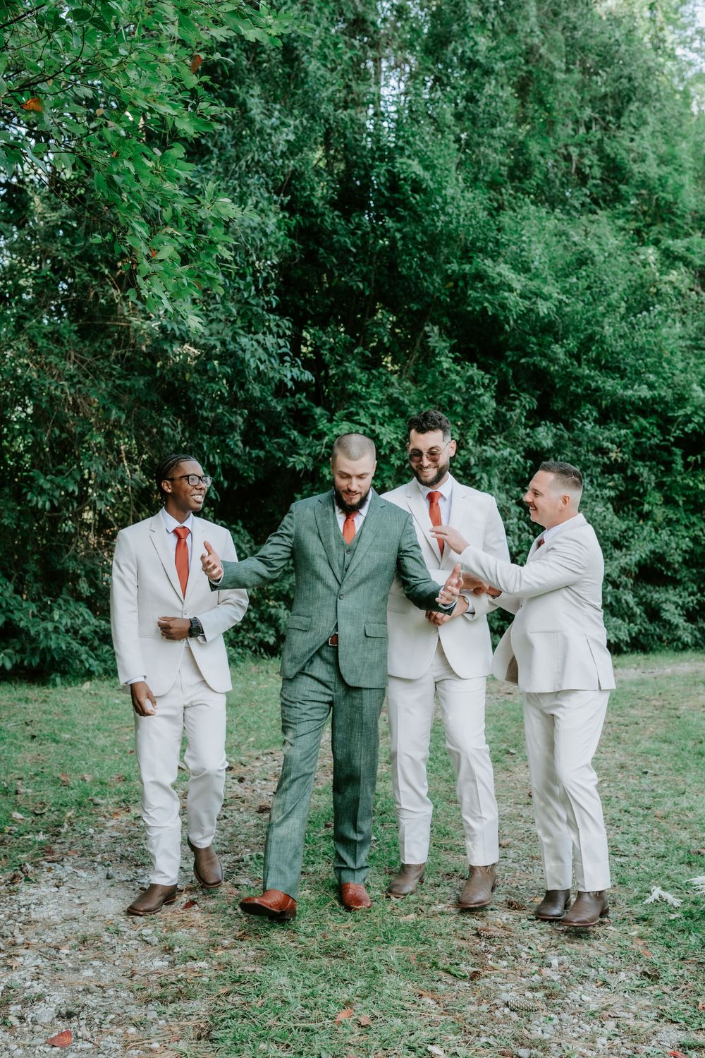 Four groomsmen in tan suits and a green suit laugh and walk together on a grassy path surrounded by trees at an outdoor wedding.