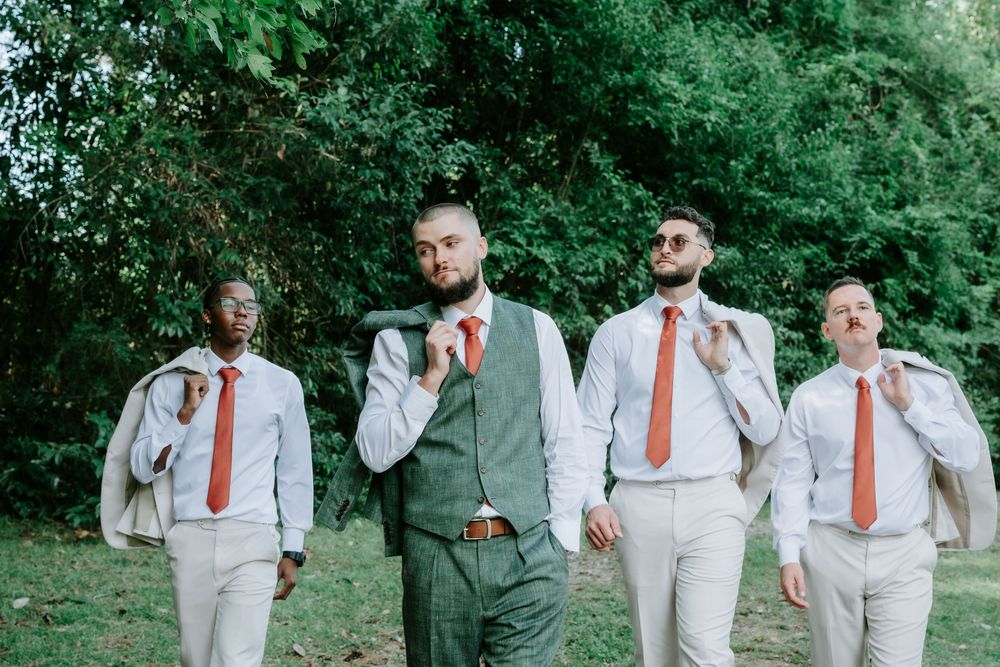 Four groomsmen in white shirts and orange ties walk together outdoors, one in a green suit and others carrying jackets over their shoulders.