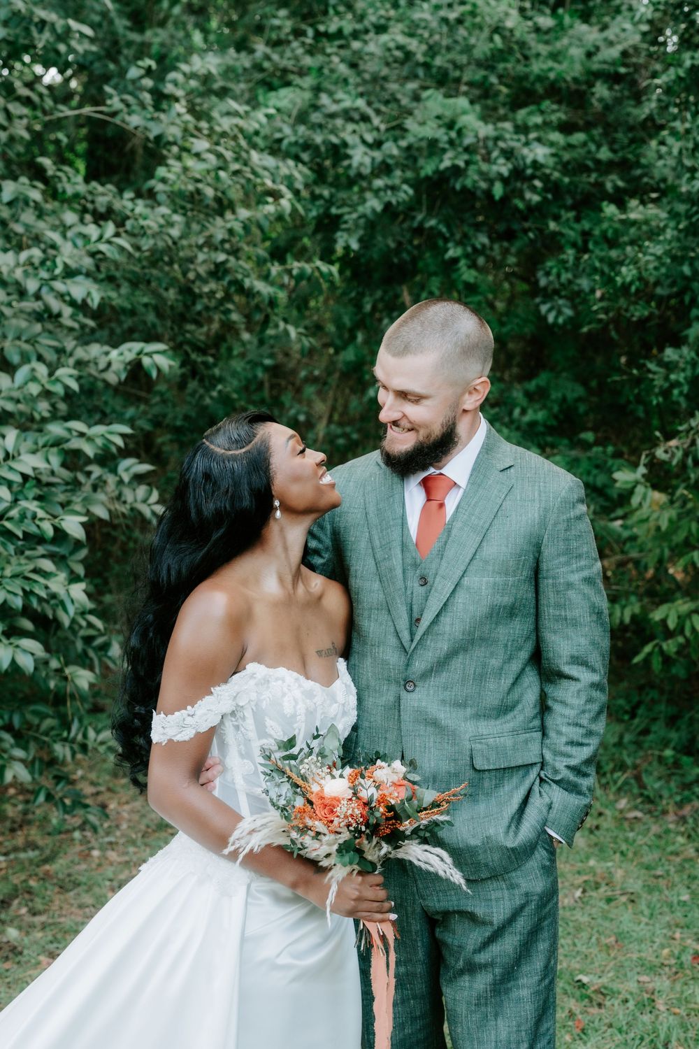 A smiling bride in an off-shoulder white gown holding a bouquet gazes up at her groom in a green suit and orange tie outdoors.