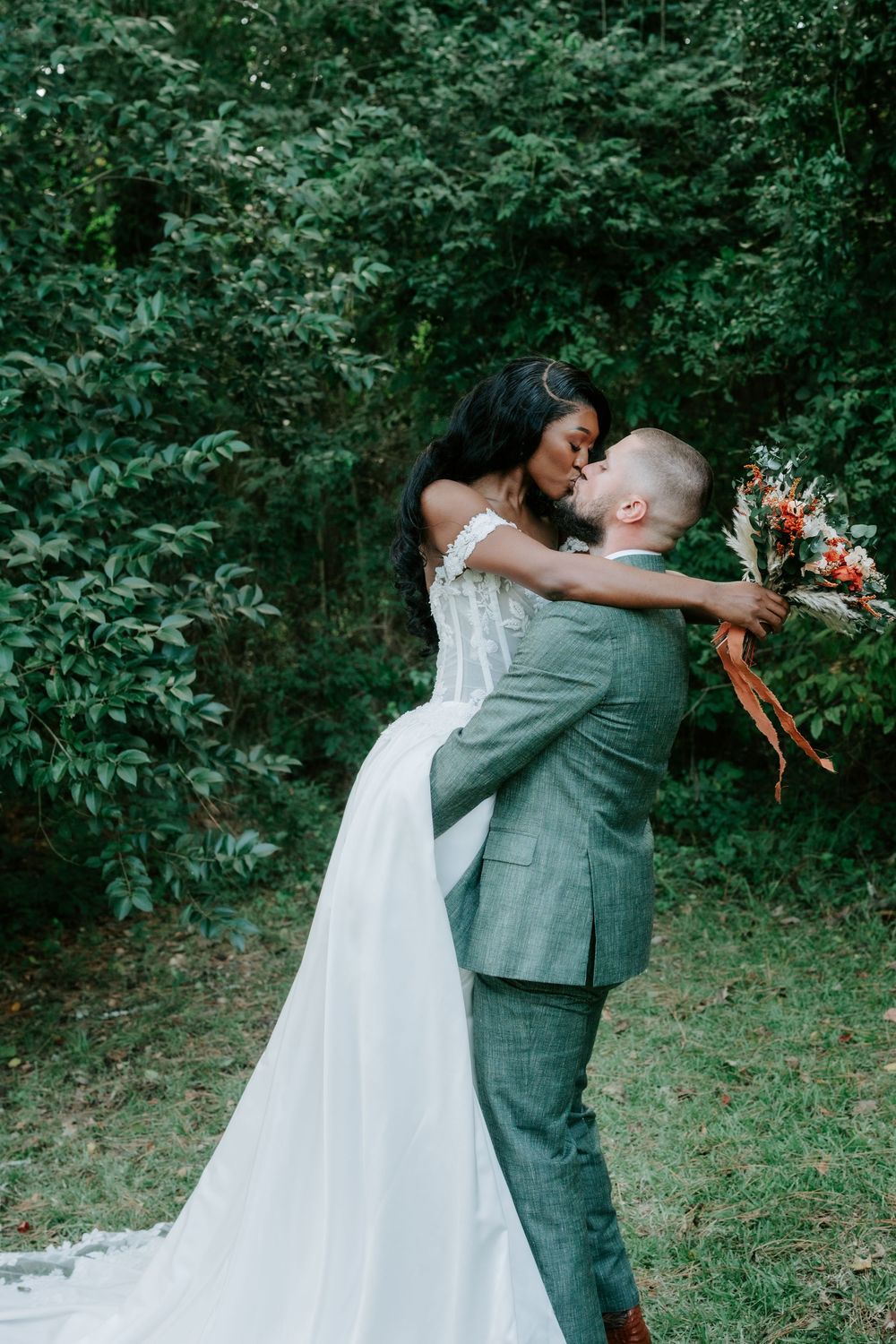 Bride in a white lace gown and groom in a gray suit share a kiss while holding a floral bouquet in a lush green forest.