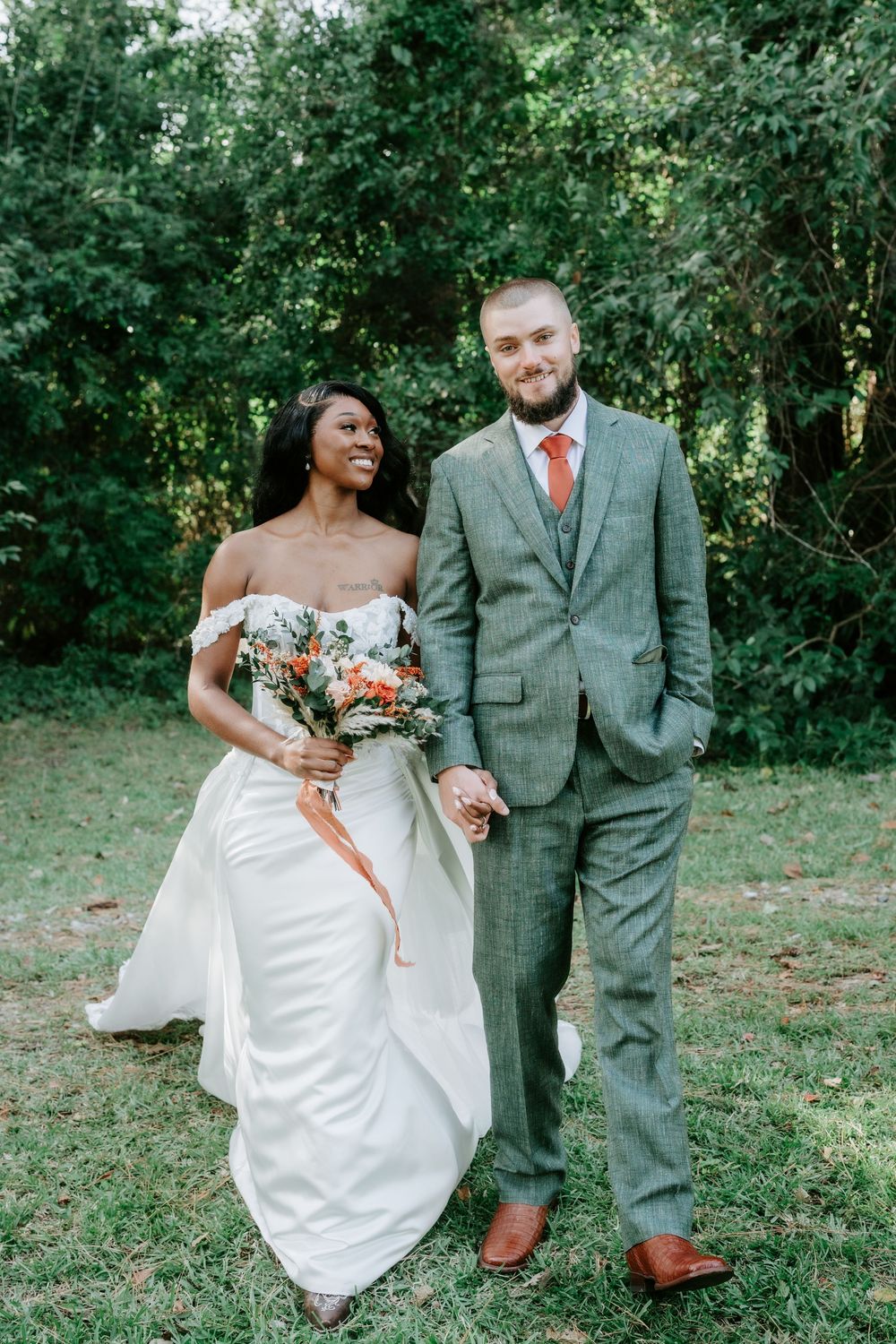 Smiling bride in an off-shoulder white gown holding a bouquet walks hand in hand with a groom in a green plaid suit outdoors.