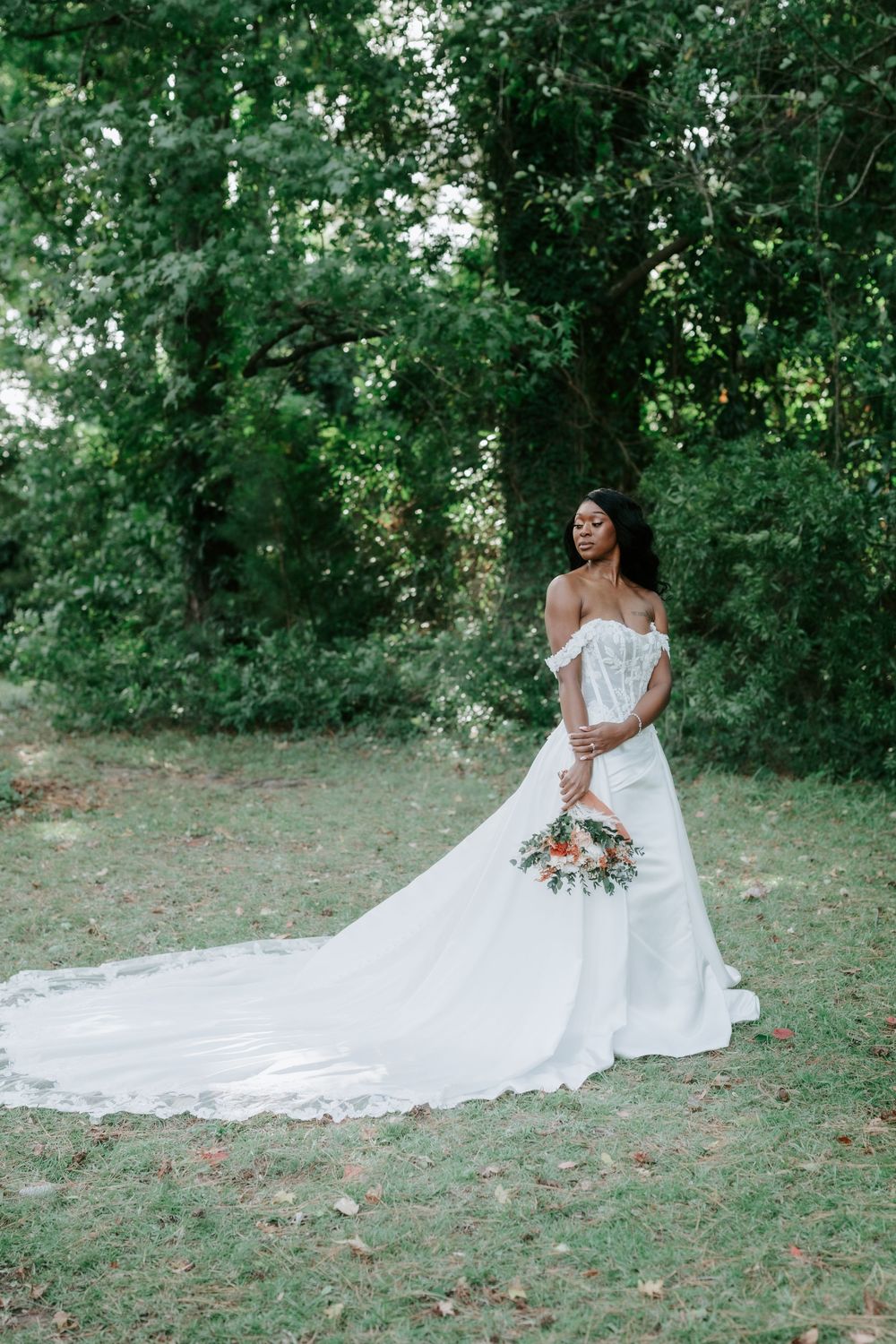 Bride in an off-the-shoulder white lace wedding gown with a long train holds a colorful bouquet while standing on a grassy lawn near trees.