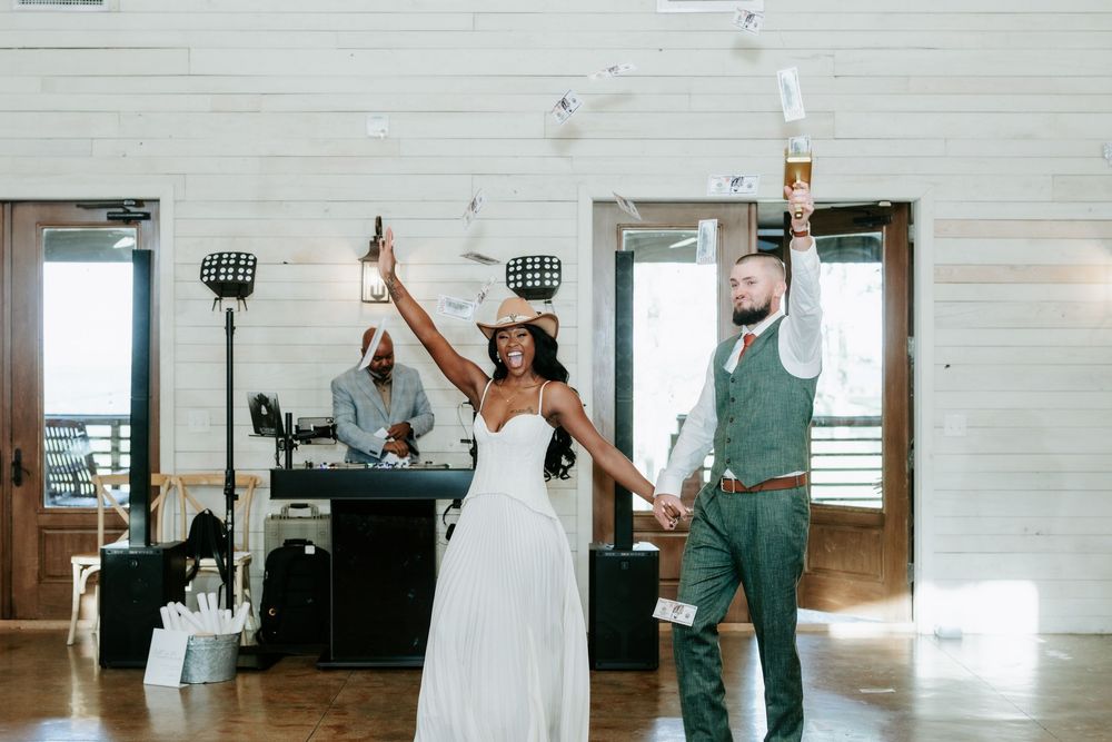 Bride in a white gown and cowboy hat and groom in a green vest raise their arms while dancing at a rustic wedding reception.