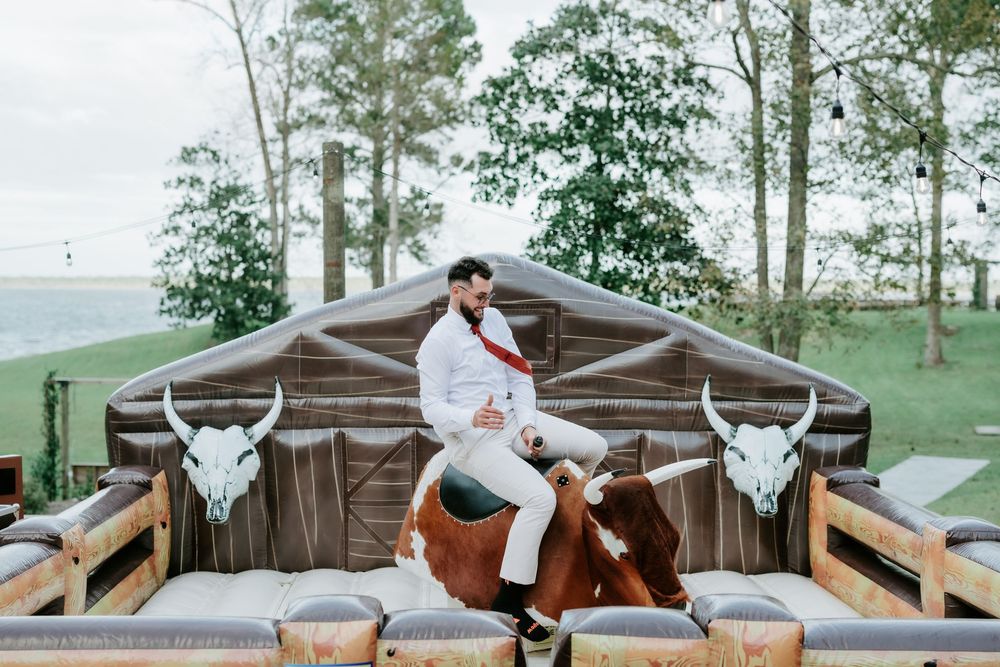Man in white shirt and red sash rides a mechanical bull at an outdoor event, surrounded by padded mats and bull-horn decorations near trees.