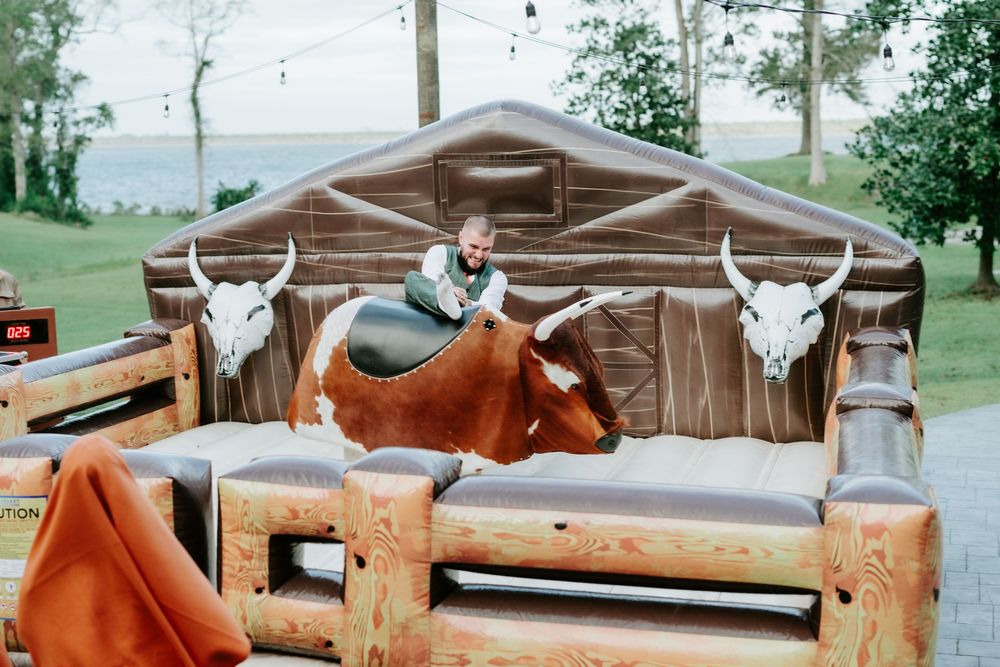 Man rides a mechanical bull at an outdoor event on a padded inflatable arena decorated with western steer skull panels near a lake.