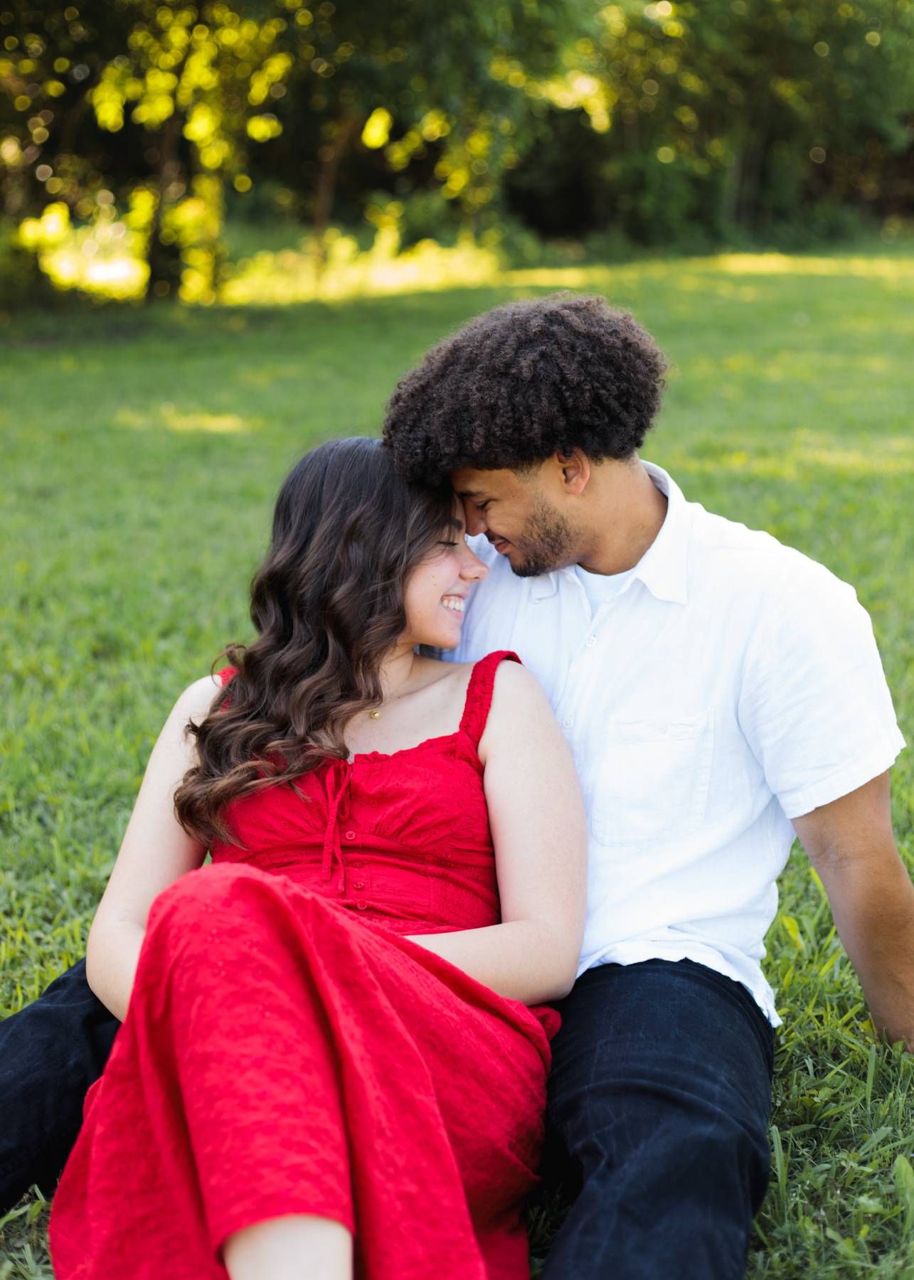 Engaged couple sits close on green grass, foreheads touching, the woman in a red dress and the man in a white shirt.