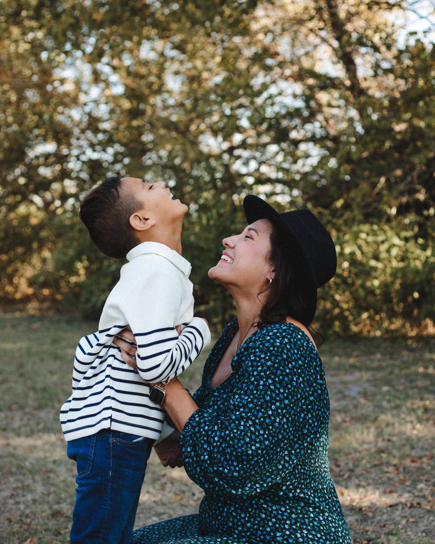 Mother in a green floral dress and black hat smiles with her young son in a striped sweater as they look up outdoors.
