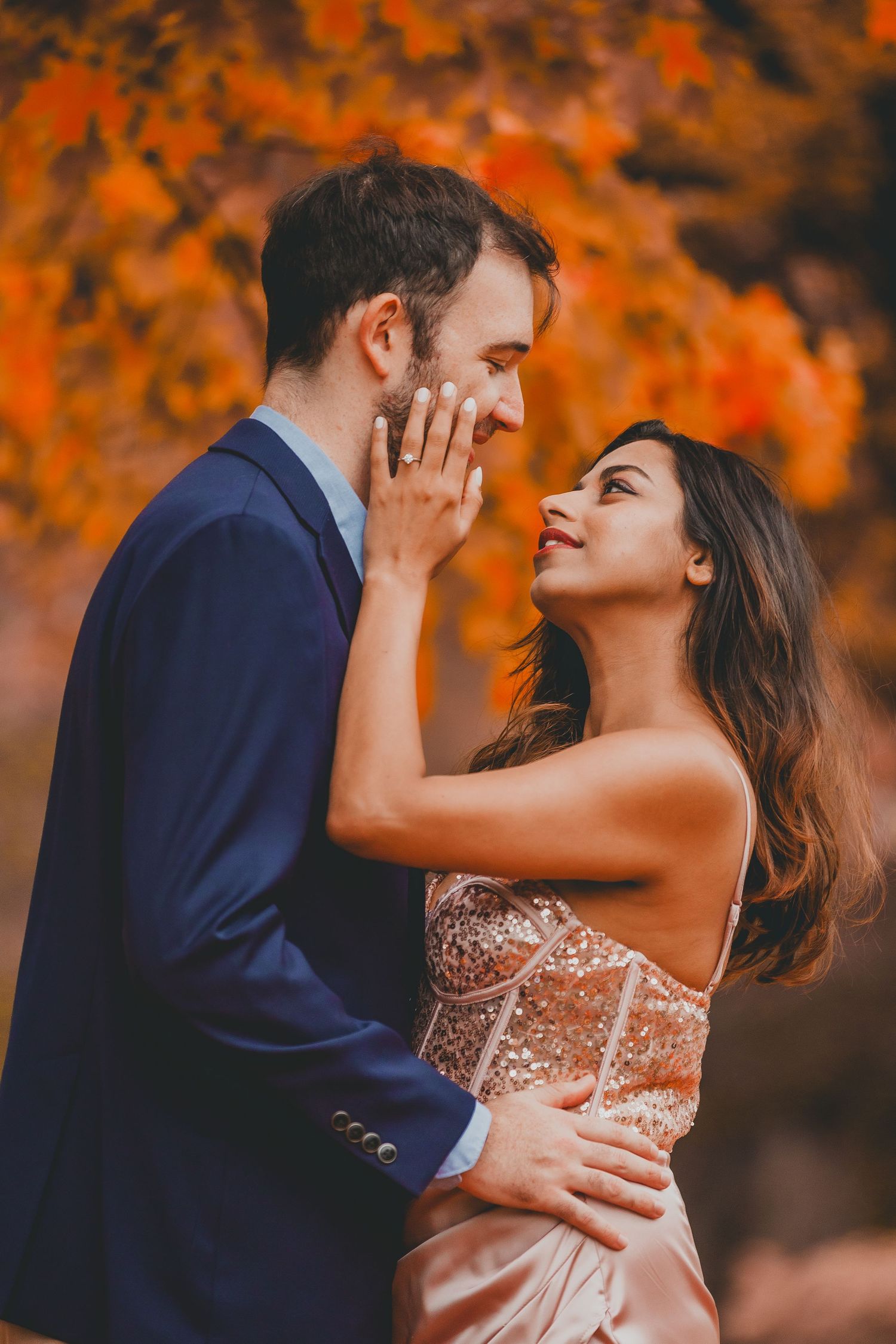 A couple embraces in an autumn setting as the woman touches the man's face, showing her ring, with warm orange foliage in the background.