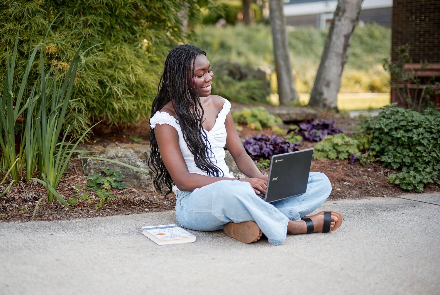 Young Black woman with braids sits cross-legged outdoors on pavement, working on a laptop in a lush garden setting.