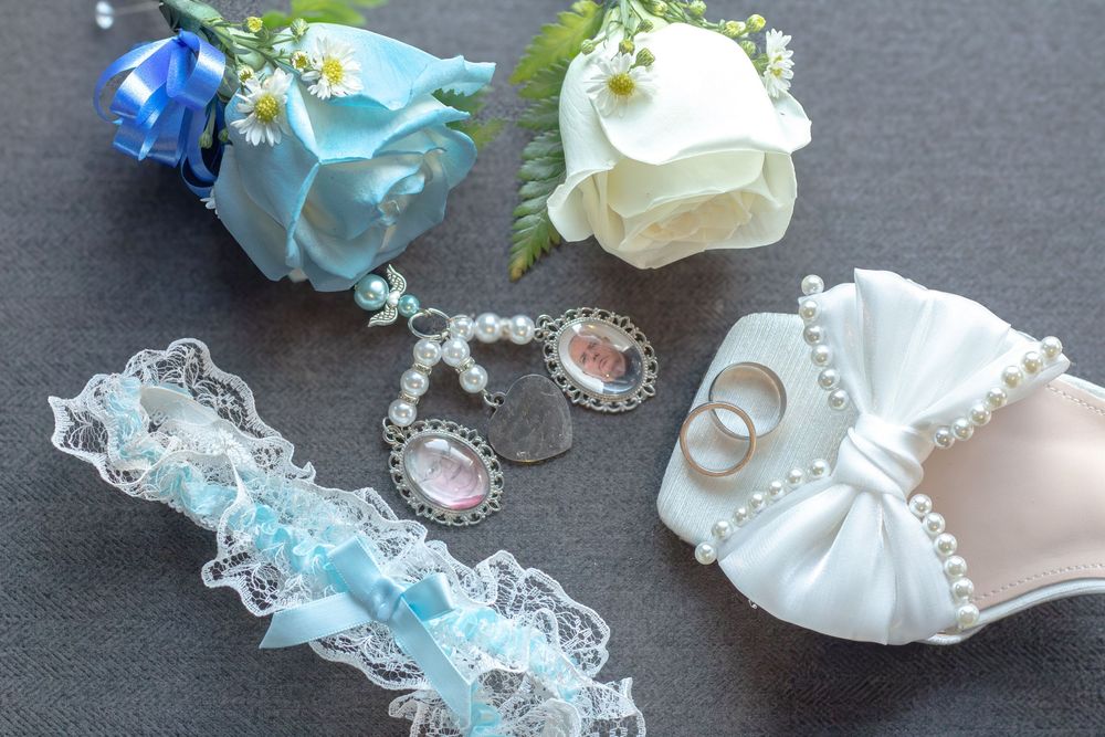 Blue and white rose boutonnieres, a lace bridal garter with blue ribbon, memory charms, and a white satin bow shoe with wedding rings.