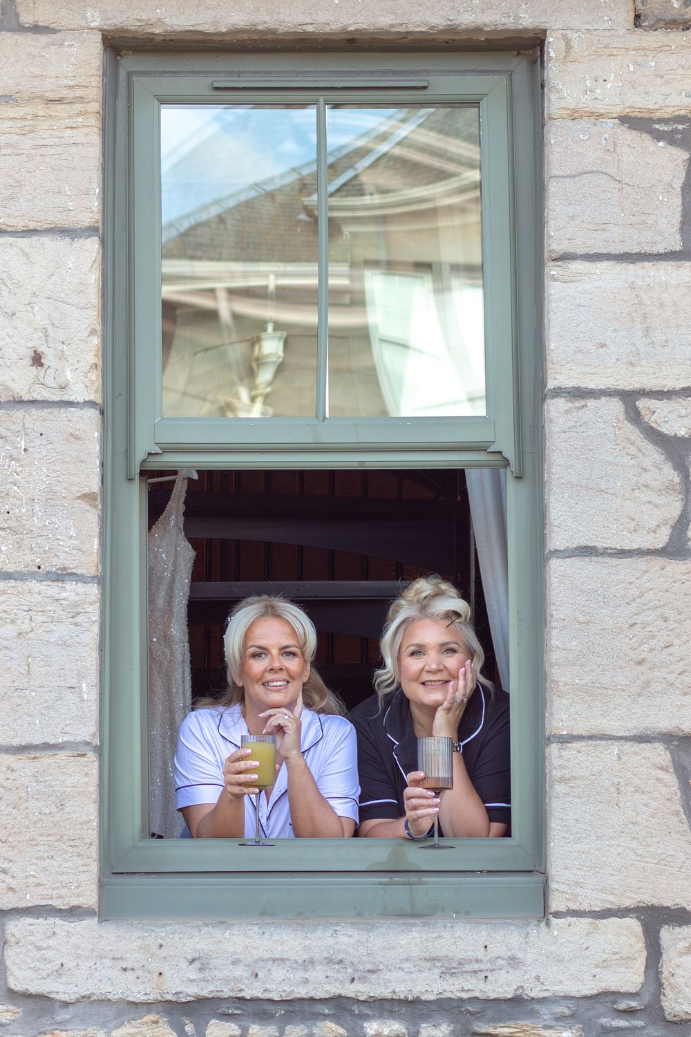 Two smiling women lean on the sill of an open green-framed window in a stone building, holding drinks and posing together.