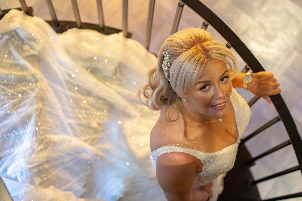 Smiling bride in an off-the-shoulder lace wedding gown with a floral hair accessory, photographed from above on a curved staircase.