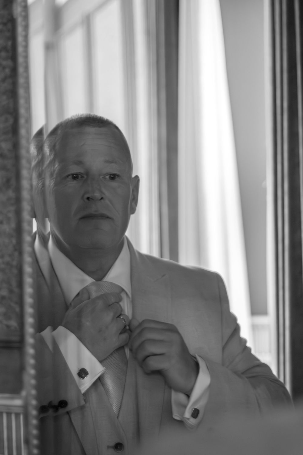 Black and white photo of a man in a suit and vest adjusting his tie in a mirror beside tall windows with sheer curtains.