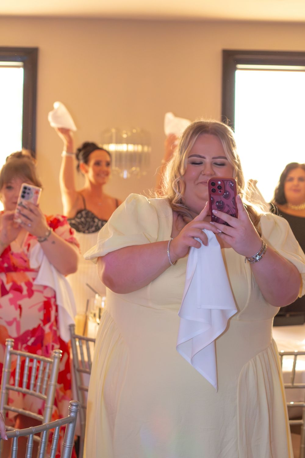 Woman in a pale yellow dress holds a white napkin while taking a photo with her phone at a festive indoor wedding reception.