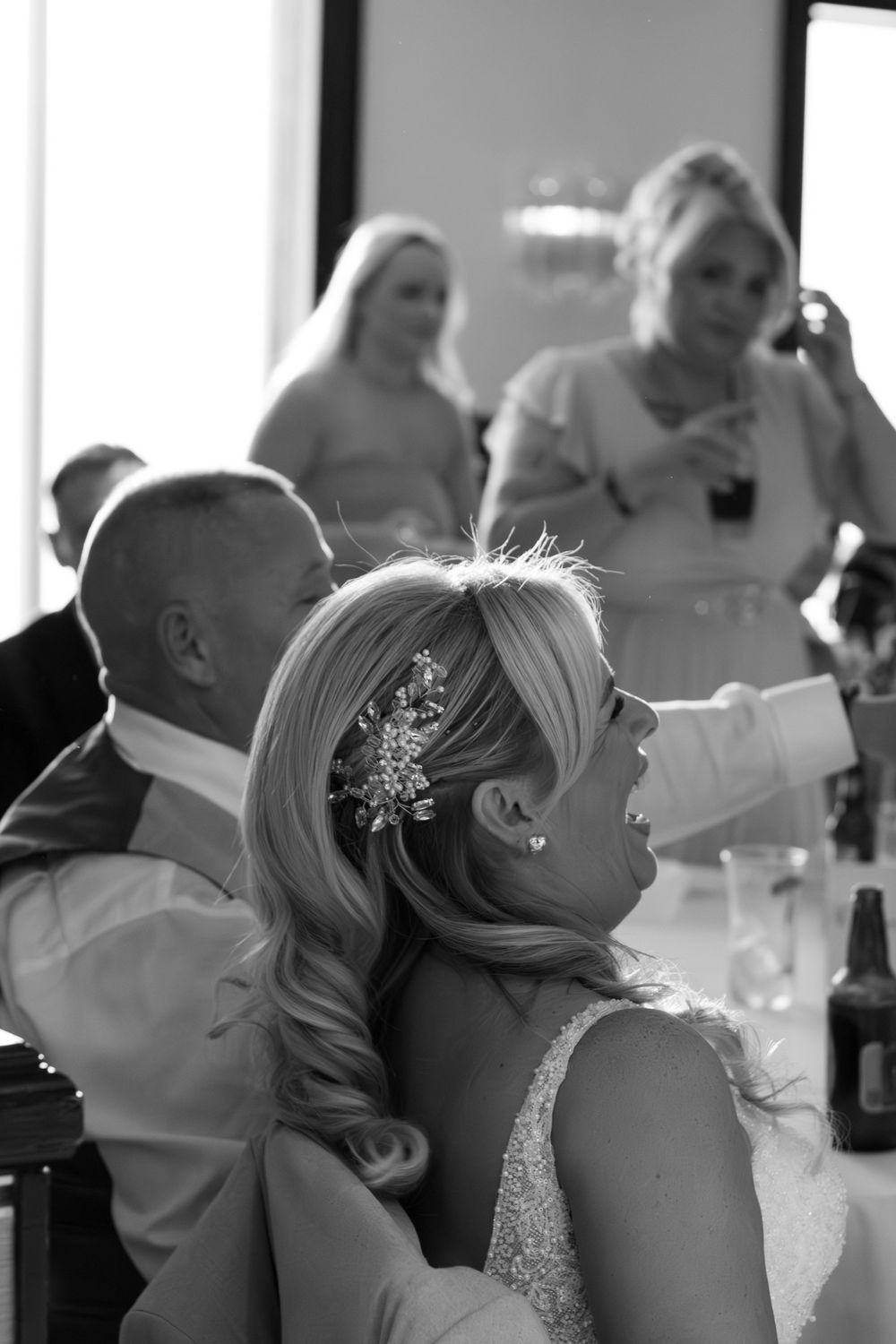 Black and white photo of a laughing bride with a floral hair clip at a wedding reception, guests standing and talking behind her.