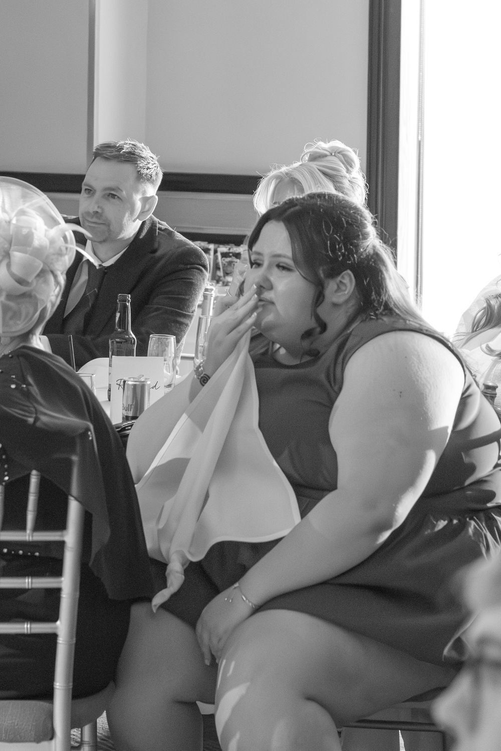 Black and white photo of a woman dabbing her eyes with a napkin while seated at a wedding reception table, with guests behind her.