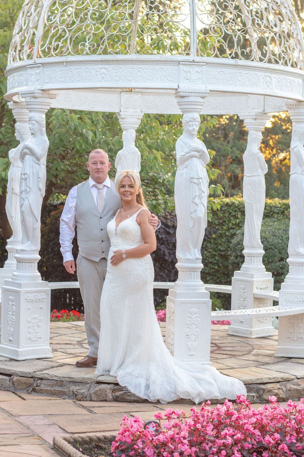 Bride in a white gown and groom in a gray vest pose together inside an ornate white gazebo with statues, beside pink flowers.