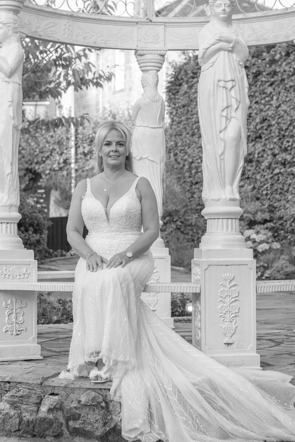 Black and white photo of a bride in a lace gown with a long train, seated on a stone ledge beneath an ornate gazebo with statue columns.