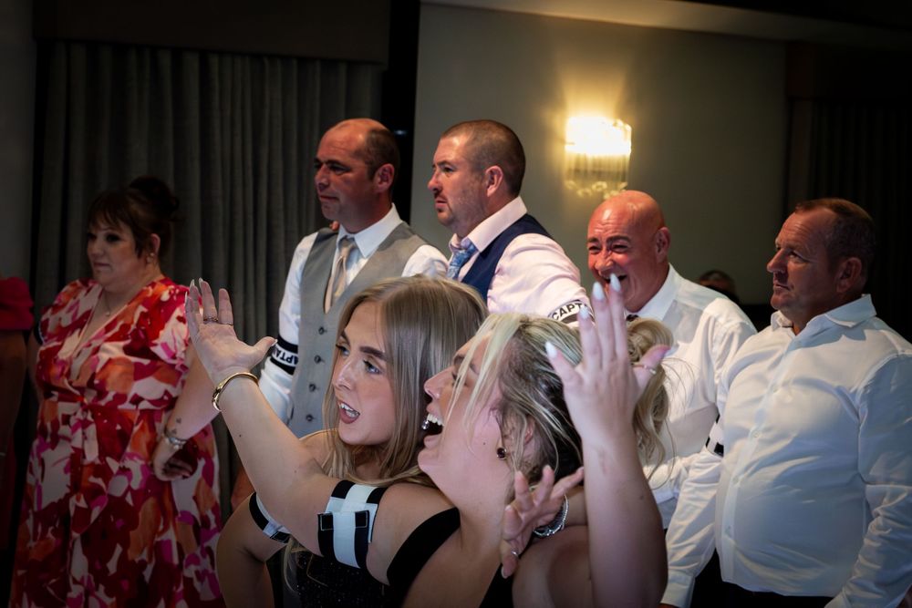 Group of smiling guests in formal attire cheering and clapping together at a lively indoor party or wedding reception celebration.
