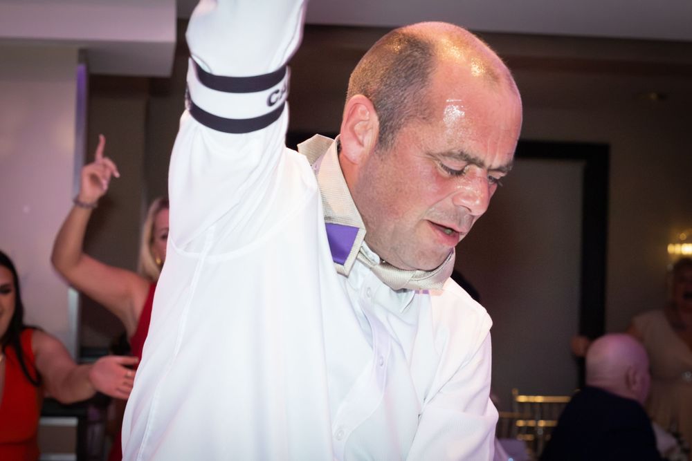 Bald man in a white shirt with a black armband dances energetically at a lively indoor party while guests celebrate in the background.