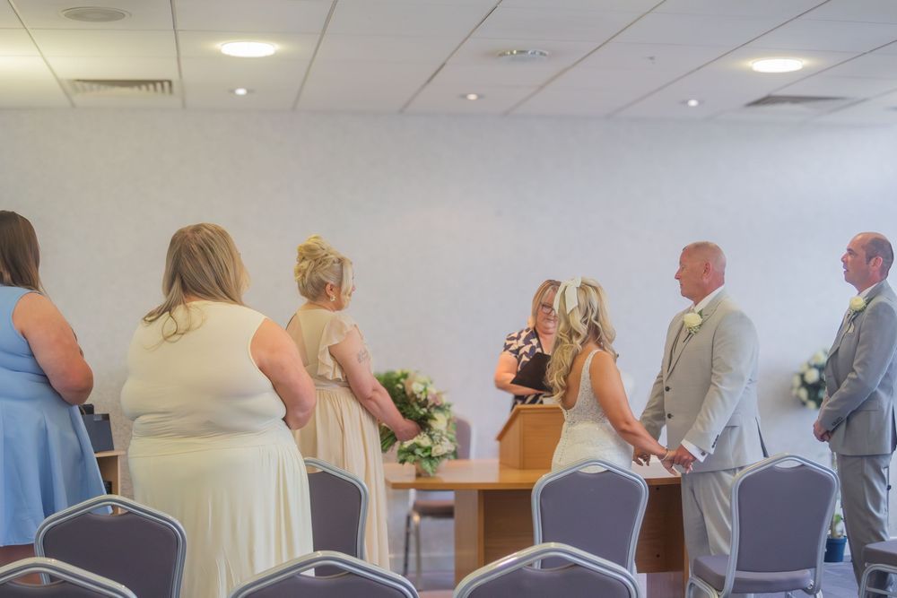 Bride and groom exchange vows at a small indoor wedding ceremony as guests in formal attire stand and watch near rows of chairs.