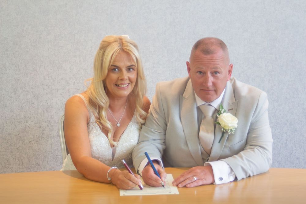 Smiling bride in a white dress and groom in a light gray suit with a white boutonniere sign wedding documents at a table.