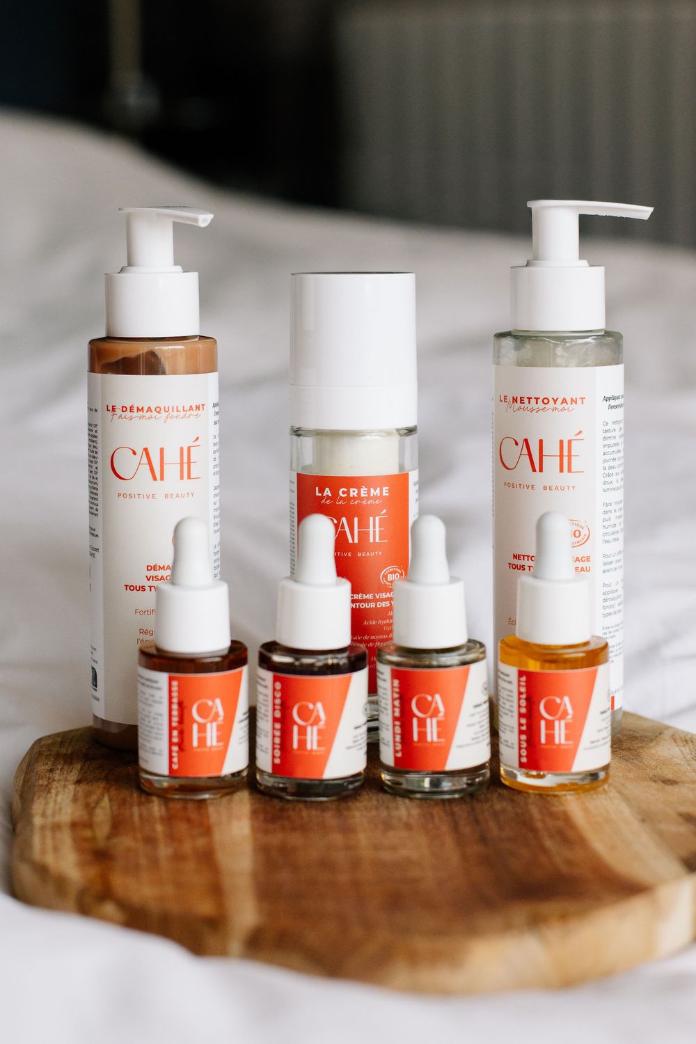 Produits cosmétiques de la marque Cahé cosmetics posés ensemble sur un plateau en bois, sur un lit aux draps blancs dans une ambiance épurée