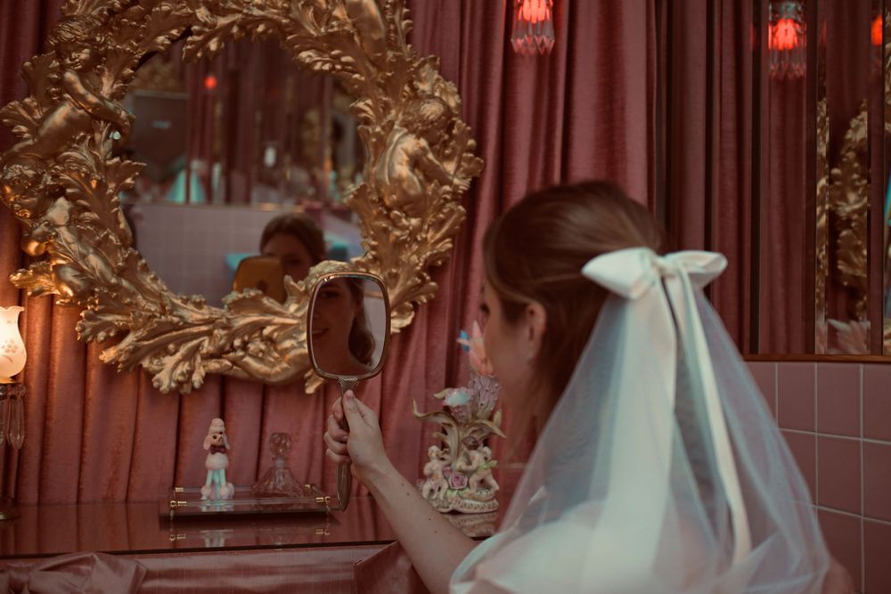 Bride in a white dress with a tulle bow veil holds a hand mirror before an ornate gold mirror in a pink-curtained room.