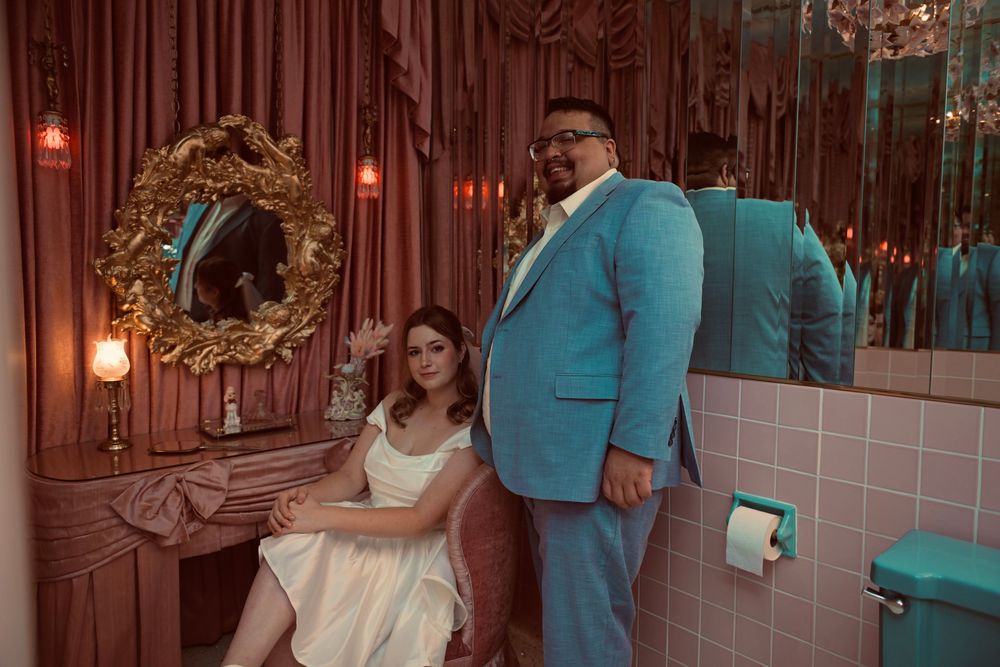 A newlywed couple poses in a retro pink-tiled lounge with a gold ornate mirror, the bride seated and the groom standing in turquoise.