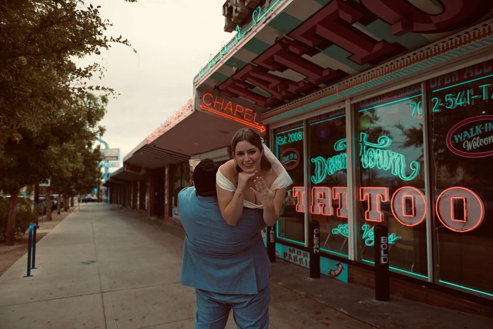 Couple embracing on a city sidewalk in front of a neon-lit tattoo shop, with the woman lifted up in a playful pose at dusk.