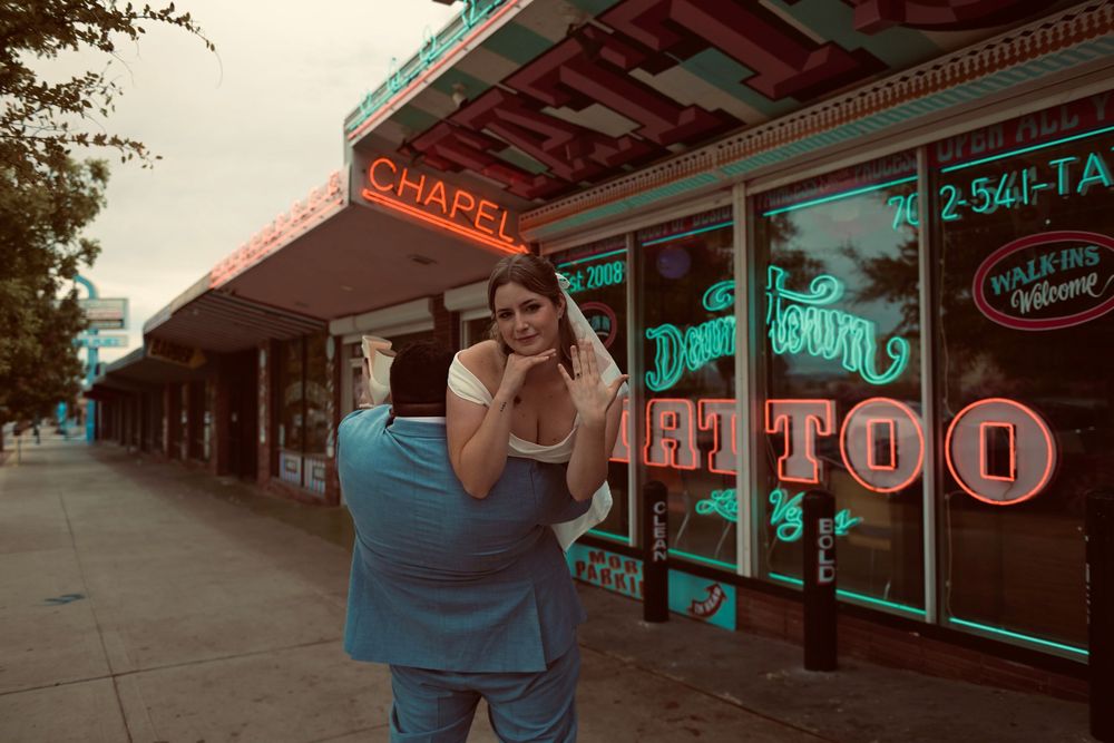 Bride in a white veil being carried by a groom in a blue suit outside a vintage wedding chapel with glowing neon signs at dusk.