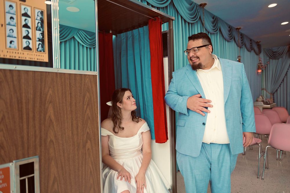 Bride in a white off-the-shoulder gown gazes up at a bearded groom in a turquoise suit inside a retro chapel with teal curtains.