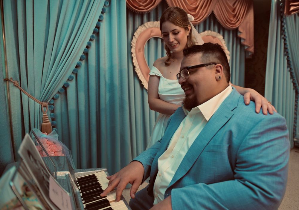 Bearded man in a teal suit and glasses plays a piano while a smiling woman in white leans on his shoulder in a vintage heart-themed room.
