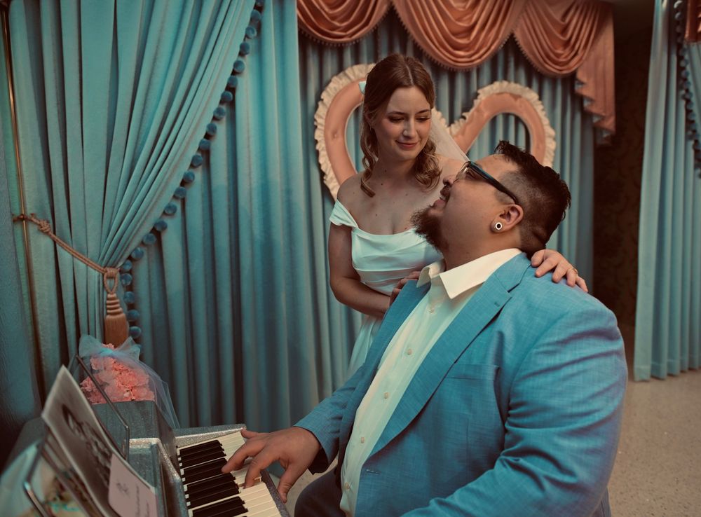 A smiling woman in a mint dress leans over a seated man in a turquoise blazer playing a piano in a teal curtained room.
