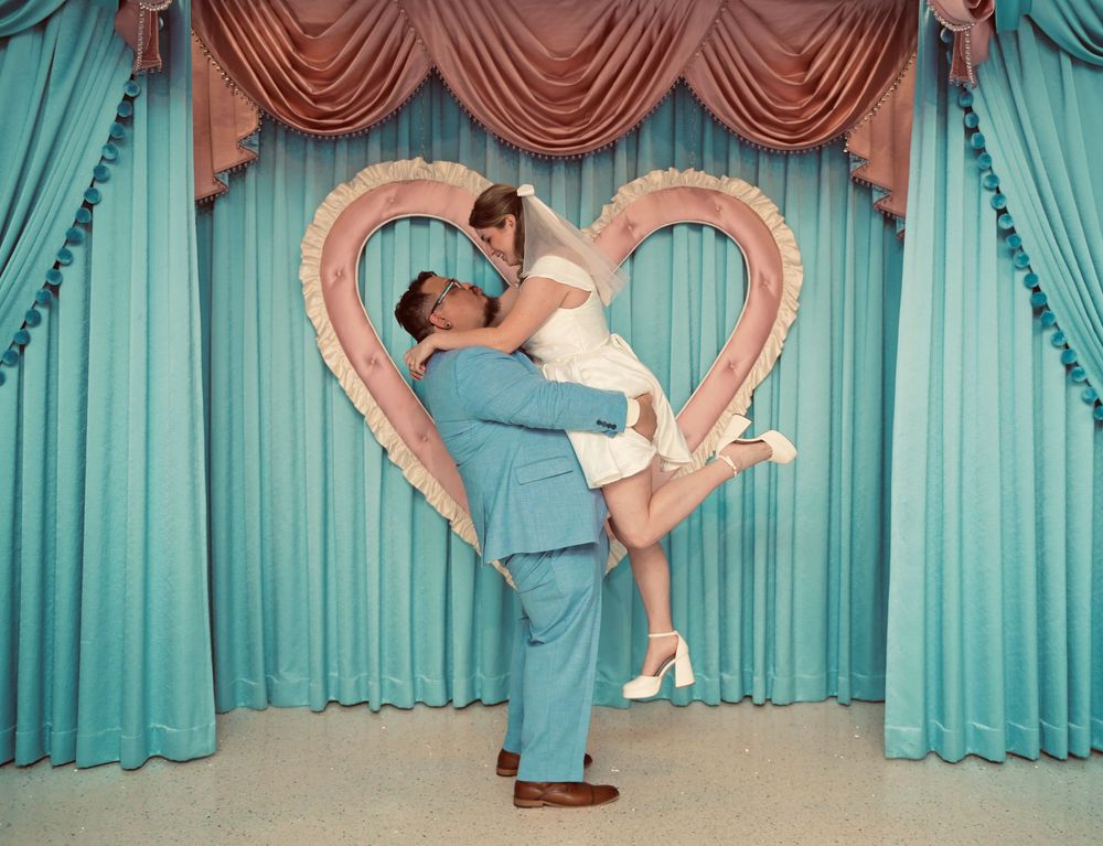 Groom in a light blue suit lifts a bride in a white dress and heels before a heart-shaped frame and teal curtains at a retro wedding chapel.